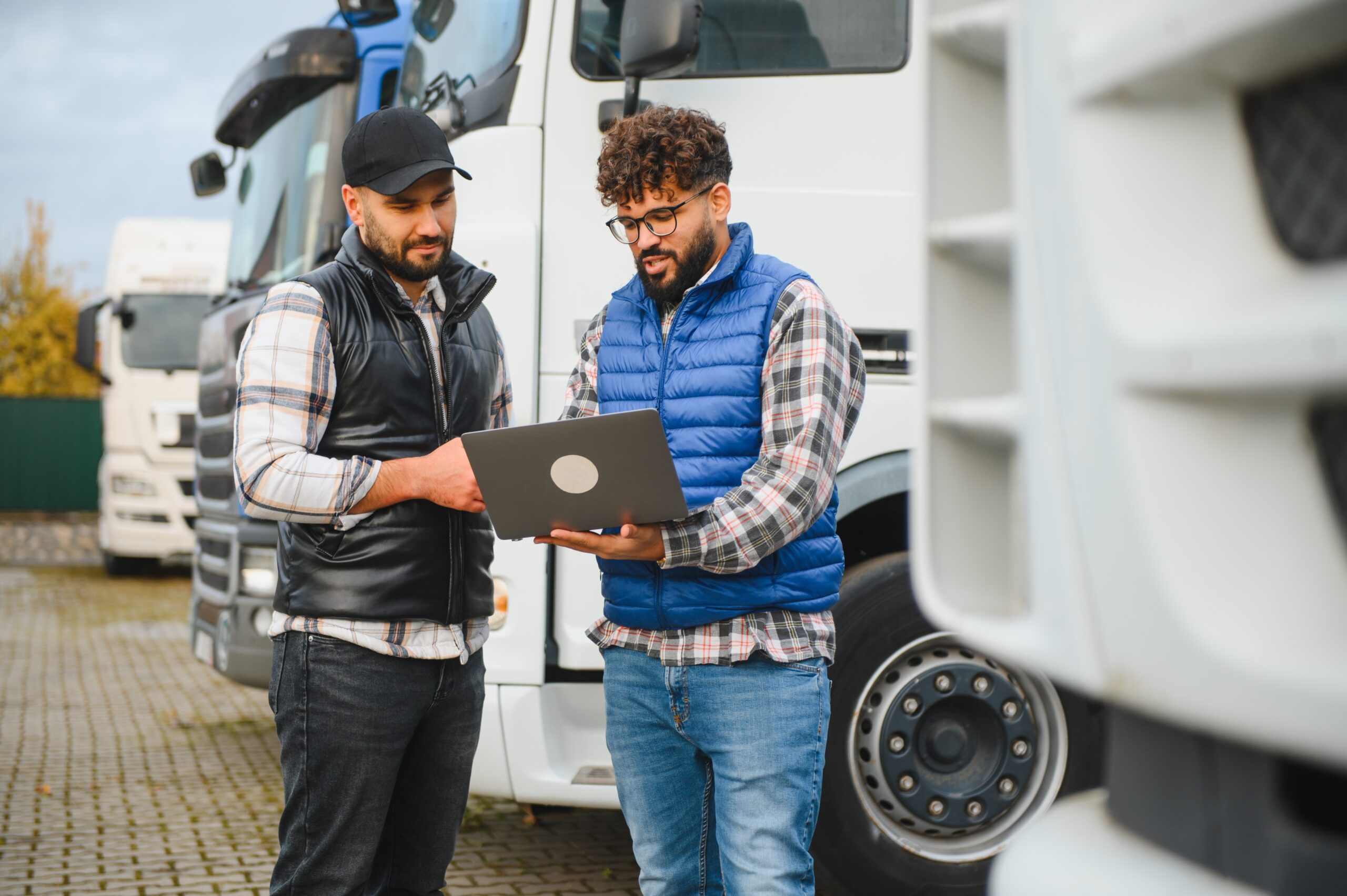 Two men stand in a logistics yard, reviewing a clipboard while discussing logistics in front of parked HGVs.